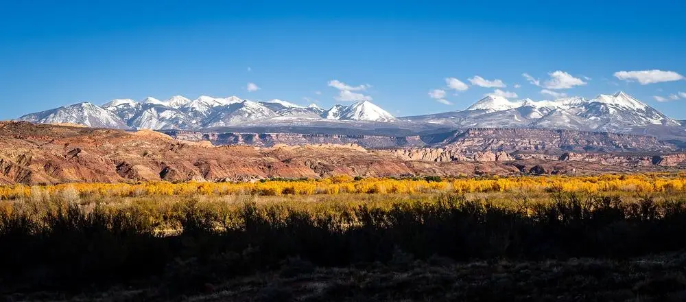 A scenic landscape featuring snow-capped mountains under a clear blue sky, with rocky red cliffs and yellow autumn trees in the foreground.