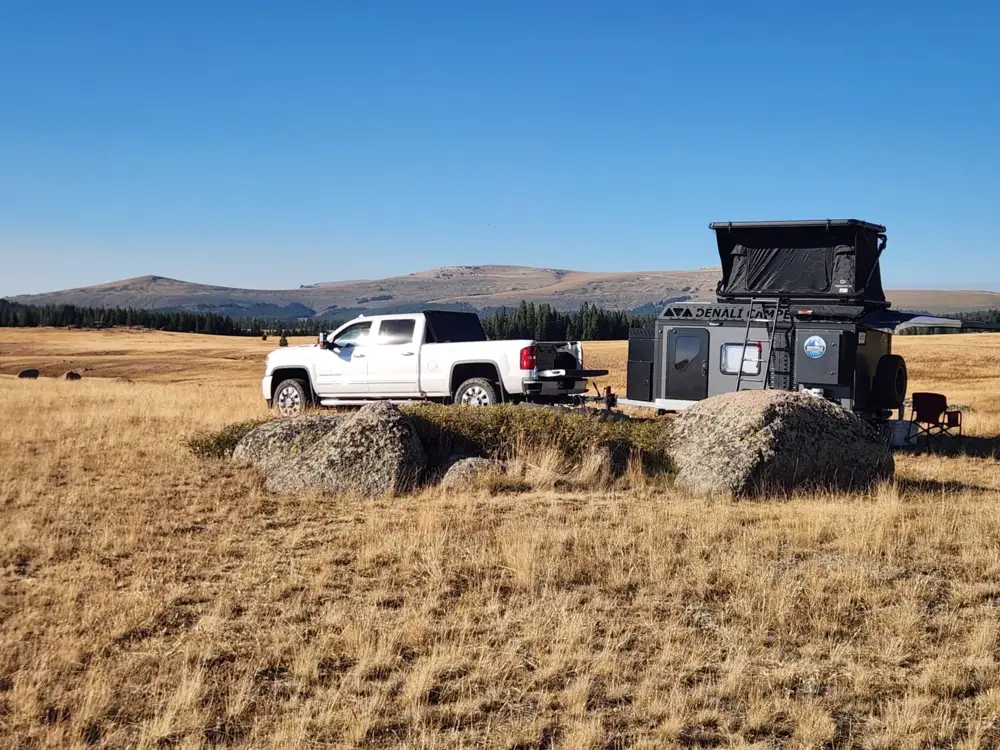 A white pickup truck is parked next to a gray camper trailer with an extended rooftop tent in a vast, dry grassland with distant hills and a clear blue sky in the background.