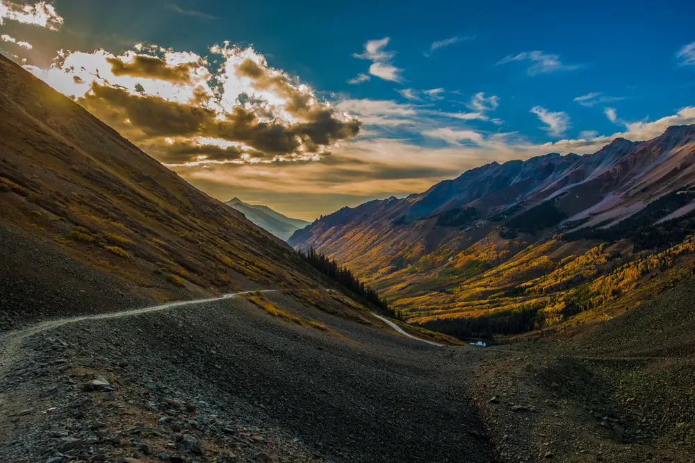 A winding dirt road cuts through a steep mountain valley, surrounded by rugged slopes and colorful autumn foliage under a dramatic sky with golden clouds at sunset.