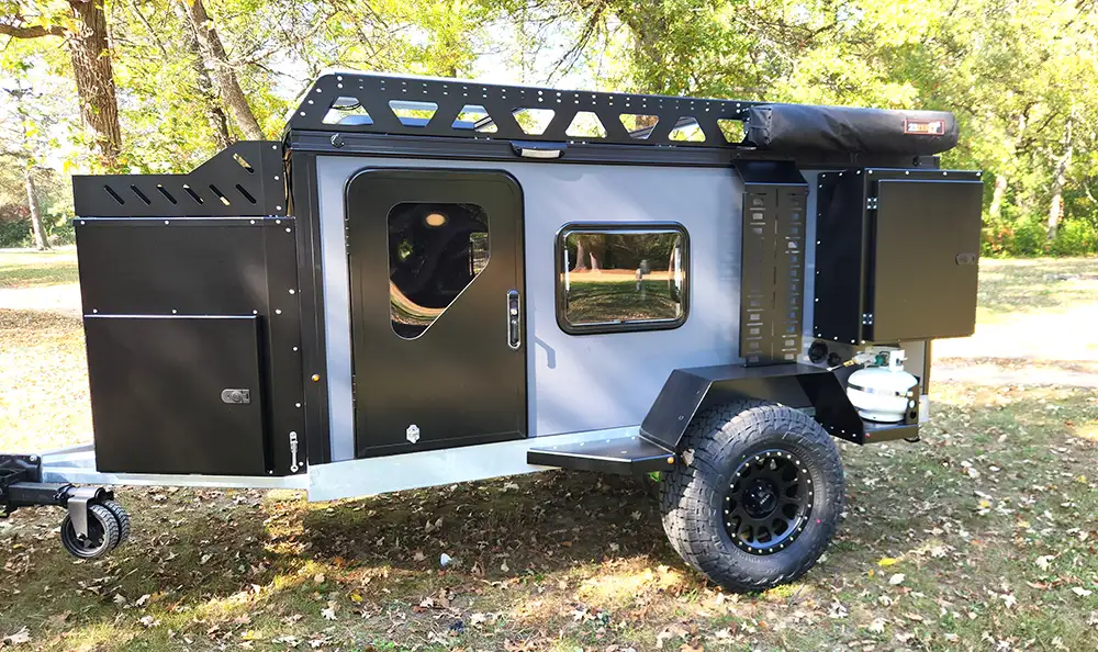 A modern black and gray off-road camping trailer with rugged tires, side compartments, a roof rack, and a small window, parked on grass among trees in a sunny outdoor setting.
