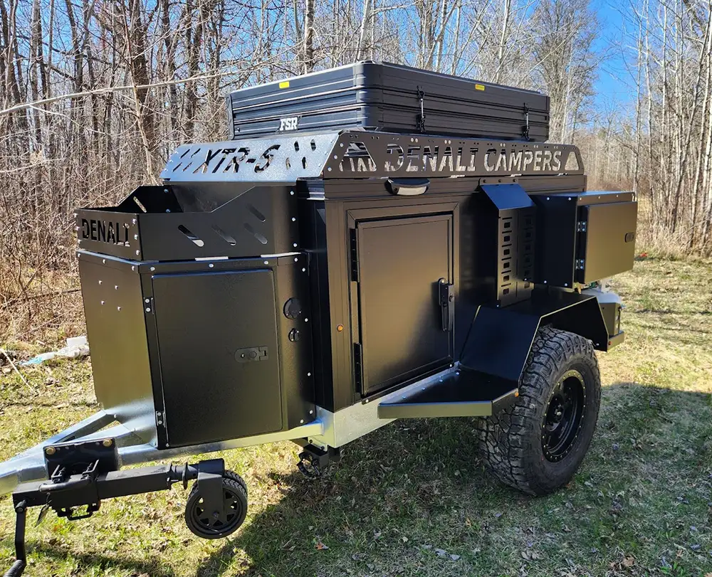 A black Denali Off-Road Camper trailer with rugged tires and a rooftop storage box is parked on grass in a wooded area with leafless trees, perfect for off-road camping as sunlight filters through.