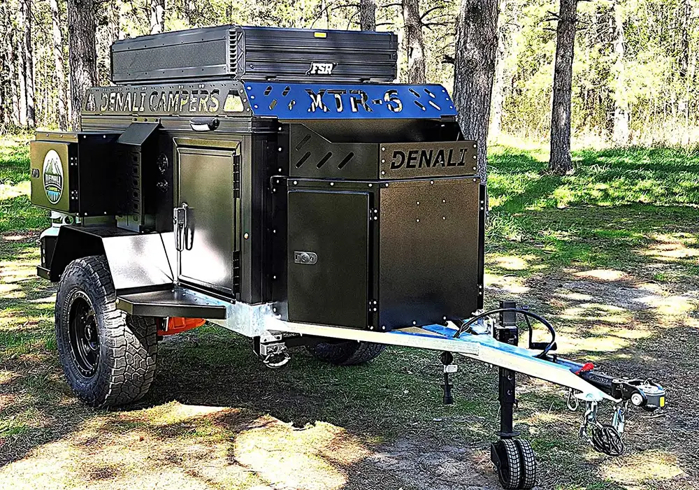 A black Denali Off-Road Camper is parked on grass among tall pine trees. The compact trailer features rugged tires, multiple storage compartments, and a long metal tow bar—perfect for adventurers seeking an expert buying guide.
