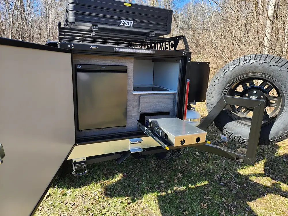 A close-up of an outdoor off-road camper kitchen setup with an open door revealing a mini fridge, storage shelves, and a slide-out stove, surrounded by grass and trees—perfect for any Off-Road Camper Guide.