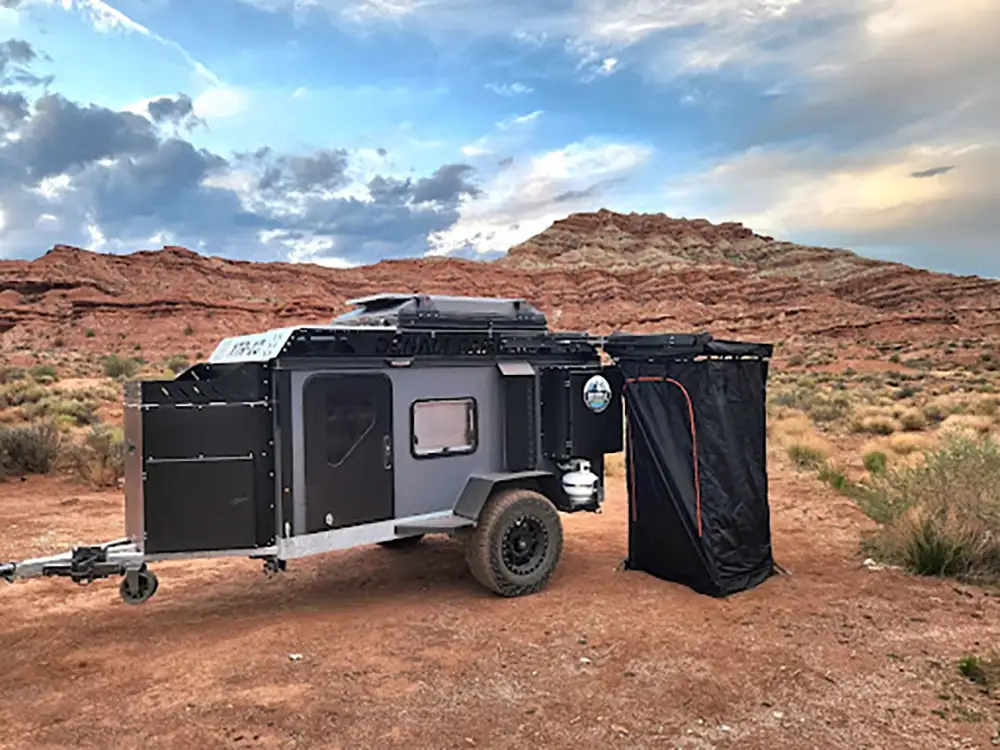 A compact off-road camper trailer with rugged tires is parked on a desert landscape of red rock formations and scattered bushes under a partly cloudy sky. A black privacy tent, perfect for any off-road camper adventure, is attached to the trailer.