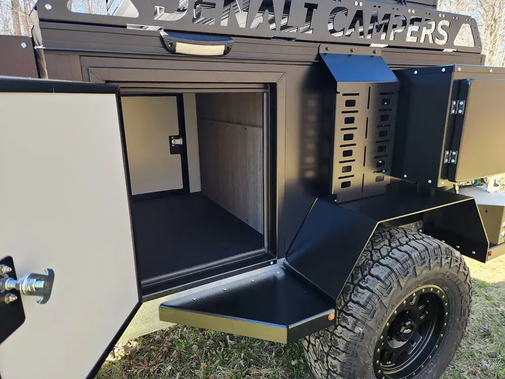 A close-up view of an off-road camping trailer with its side door open, revealing a spacious storage compartment. The trailer has rugged tires, black metal exterior, and "DENALI CAMPERS" signage on top.