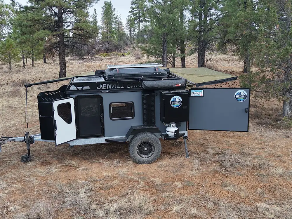 A rugged off-road camper with large tires, rooftop storage, and an open rear hatch sits in a dry pine forest clearing. The trailer, perfect for off-road camping, features adventure decals and attached camping gear.