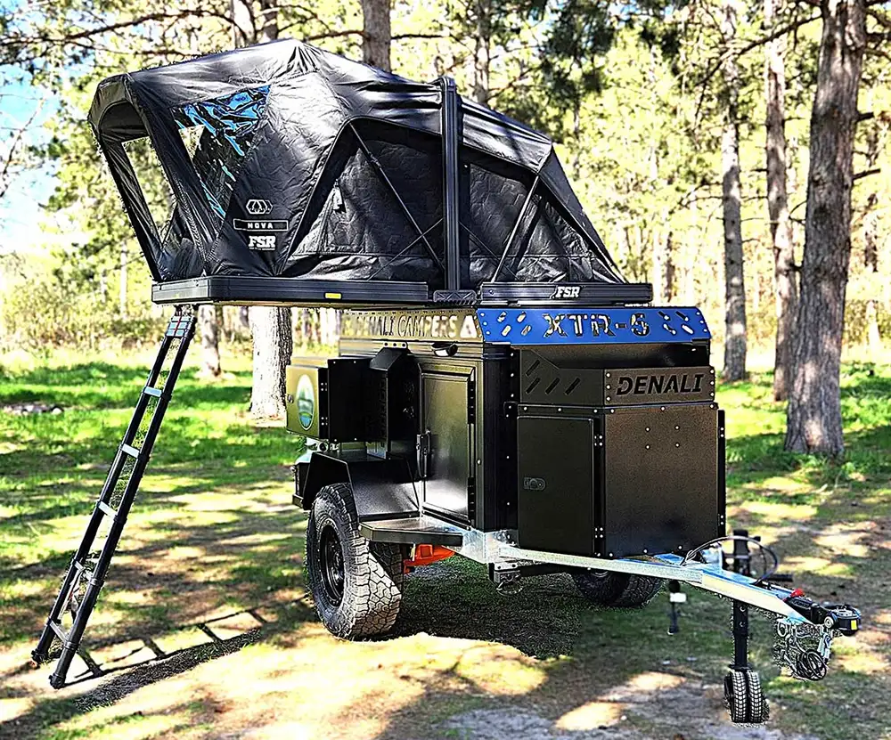 A rugged off-road camper with large tires, a blue and black metal body, and a rooftop tent with ladder is set up in a wooded area on a sunny day—perfect for your next off-road camping adventure.