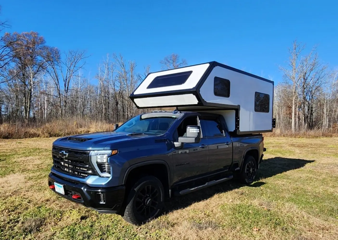 A dark blue pickup truck with a white camper attached to its bed is parked on grass. The sky is clear and blue, and leafless trees are visible in the background.