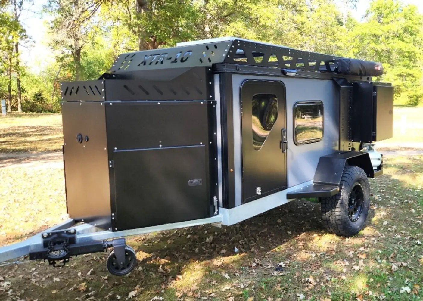 A rugged, black and silver off-road camping trailer with large tires stands parked on grass in a wooded area, featuring roof storage, side doors, and spacious trailers storage compartments.