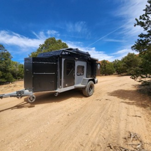 A gray and black off-road camper trailer parked on a dirt road, surrounded by trees and under a bright blue sky with wispy clouds.