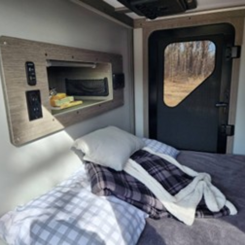 A cozy bed with plaid and checkered bedding is inside a small camper or RV. A window and a shelf with toiletries are on the wall, and a black door lets in sunlight from outside.