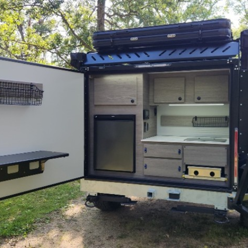 A compact camper trailer kitchen with wood cabinets, a mini fridge, sink, and stove is shown outdoors. The open door has storage shelves and a fold-down table. Trees and greenery are visible in the background.