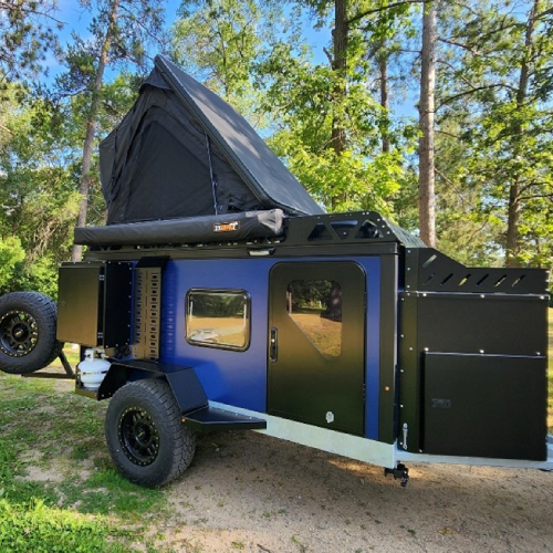 A blue off-road camping trailer with black accents is parked outdoors among trees. It features an open rooftop tent, spare tire, storage compartments, and rugged tires, ready for outdoor adventure.