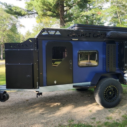 A blue and black off-road camping trailer with large tires, a metal roof rack, and side storage compartments is parked on a dirt area surrounded by trees and greenery.