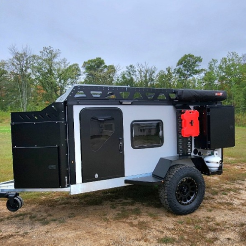 A compact off-road camper trailer with black and white exterior, rugged tires, rooftop rack, red fuel canister, and side storage compartments, parked on a grassy area with trees in the background.
