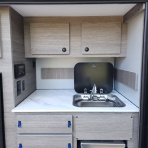 A compact kitchenette with wood-tone cabinets, a stainless steel sink with a faucet, white countertop, and drawers, set against a light-colored backsplash inside a small space.