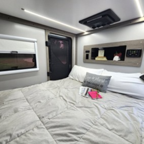 A cozy RV bed with a light gray comforter, white pillows, and a decorative pillow that reads “Give Up The Ghost.” Books and a pink tablet rest on the bed; a window, door, and built-in shelf are in the background.