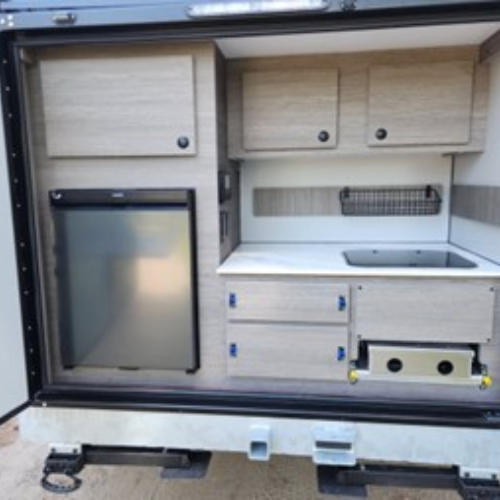 A compact outdoor kitchenette inside a trailer features a mini fridge, cabinets, drawers, a small sink, and a pull-out stovetop, all in a light wood finish.