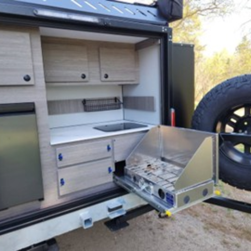 A compact outdoor camper kitchen features a stove, sink, countertop, drawers, cupboards, small fridge, and a metal pull-out two-burner cooktop, all neatly built into the side of a trailer.