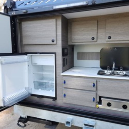 Outdoor camper kitchen with open mini-fridge, sink, two-burner stovetop, cabinets, and drawers. The kitchen is built into a vehicle and features wood-look paneling and compact appliances.