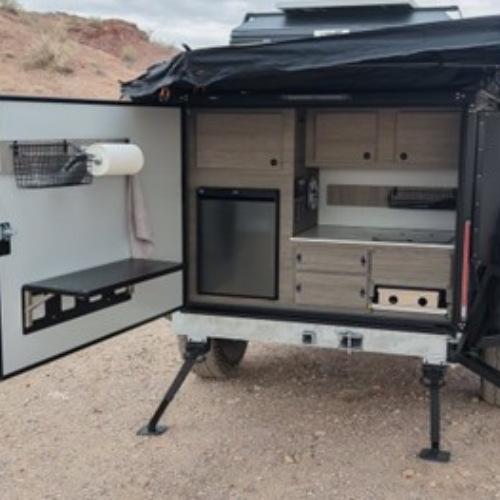 A compact outdoor camper kitchen features cabinets, a mini fridge, a sink, fold-out counter space, a paper towel holder, and storage compartments, set up on rocky terrain.