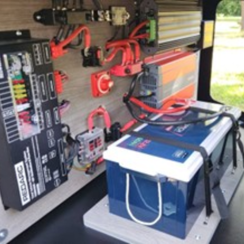 A close-up of an electrical setup featuring a deep cycle battery, inverter, fuse box, and various wiring components installed on a wooden panel, likely for an off-grid power system or camper van.