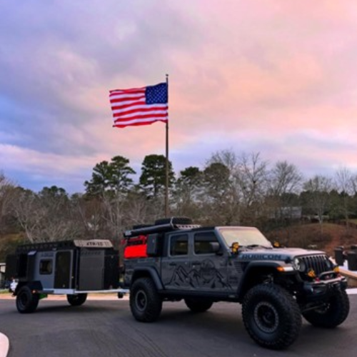 A black off-road vehicle towing a camping trailer is parked on a road at sunset, with a large American flag flying on a pole and trees in the background.