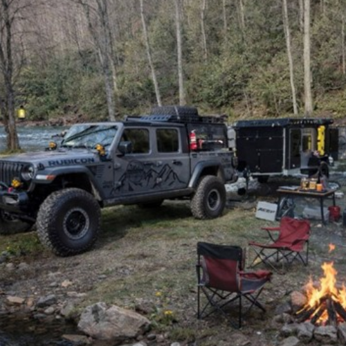 A gray off-road Jeep with a trailer is parked beside a river in a wooded area. Camping gear, two red chairs, a table, and a campfire are set up nearby. Trees and flowing water are visible in the background.
