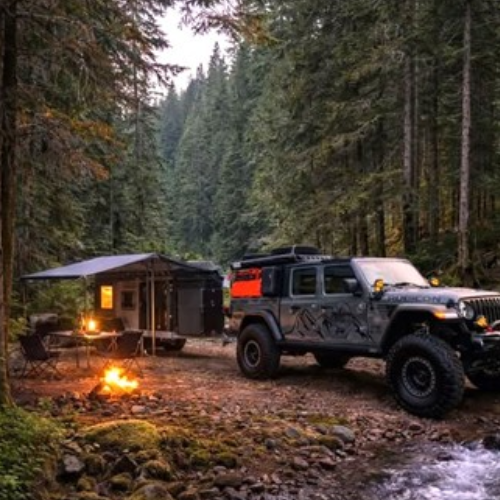 A rugged off-road vehicle and camper trailer are parked by a campfire in a forest clearing, surrounded by tall evergreen trees. Camping chairs and an awning are set up next to the trailer beside a shallow creek.
