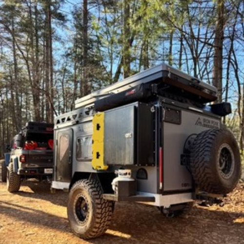 An off-road vehicle with a camping trailer is parked on a dirt path in a forested area, surrounded by tall trees and sunlight filtering through the branches.
