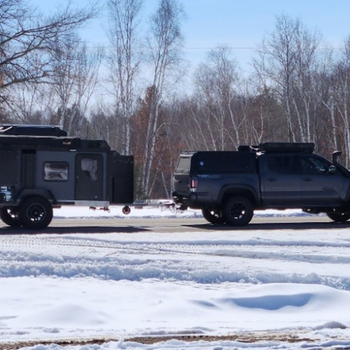 A black off-road truck with a matching black camper trailer is parked on a snow-covered lot, surrounded by leafless trees under a clear blue sky.