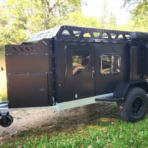 A black off-road camping trailer with rugged tires, side door, small window, and storage compartments is parked on grass with trees in the background.