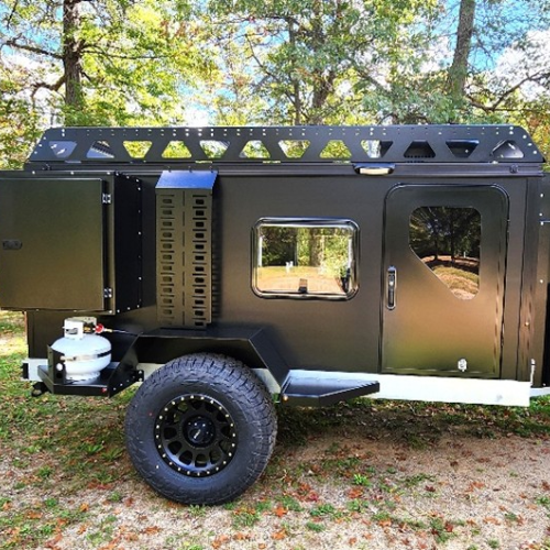 A rugged black off-road camping trailer with large tires, a roof rack, and an attached propane tank is parked on grass amid trees in a wooded area.