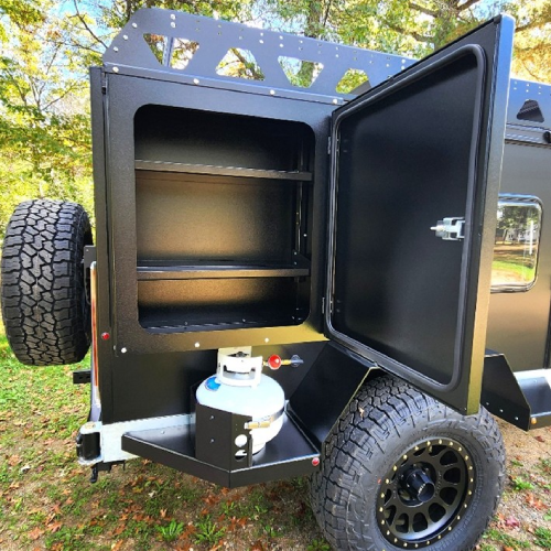 A black off-road trailer with an open side compartment showing empty shelves and a mounted propane tank below, parked on grass with trees in the background.
