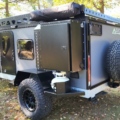 A rugged off-road camping trailer with large tires, a mounted spare tire, a propane tank, storage compartments, and a black rooftop cargo bag, parked on grass among trees.