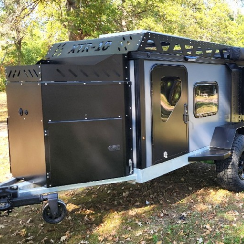 A black off-road camping trailer with rugged tires and a roof rack is parked on grass, surrounded by trees. The trailer has a rectangular front section and side windows, designed for outdoor adventures.