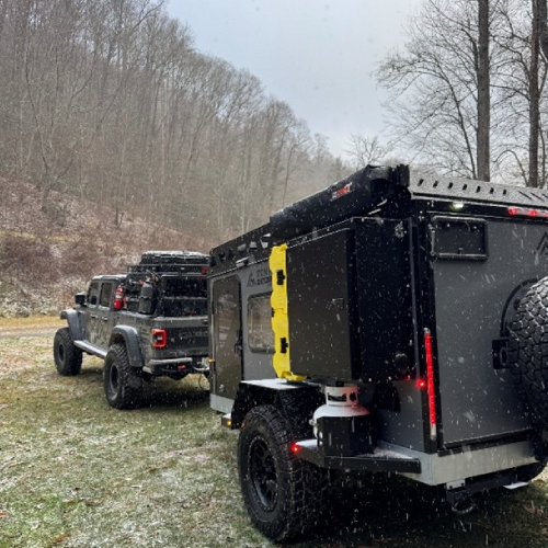 A rugged off-road vehicle towing a black adventure trailer with a yellow fuel can, parked on grass with light snowfall and bare trees in a hilly, wooded area.