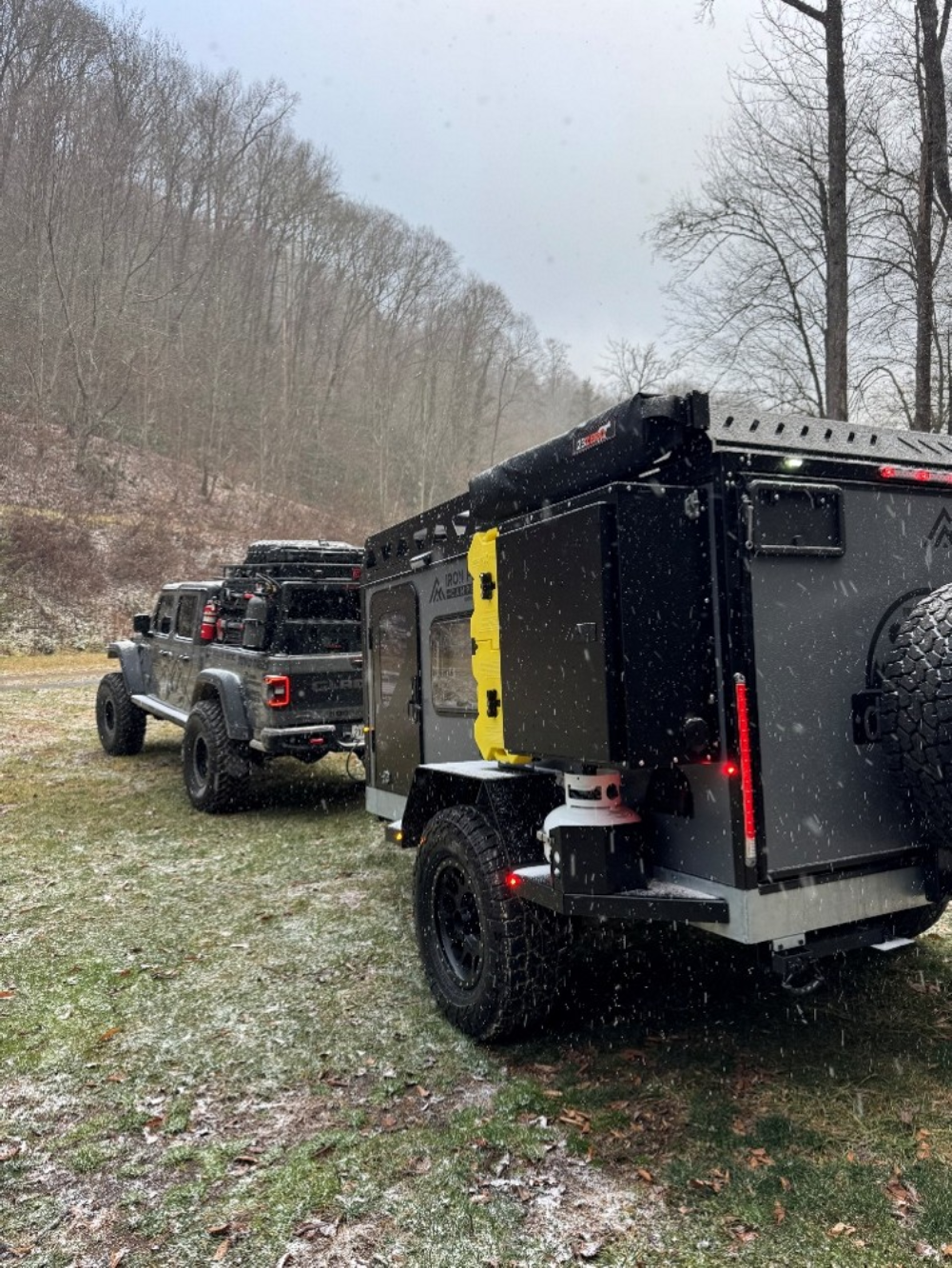 A black off-road vehicle and an attached camper trailer are parked on a grassy area with light snow falling, surrounded by leafless trees and hills in the background.