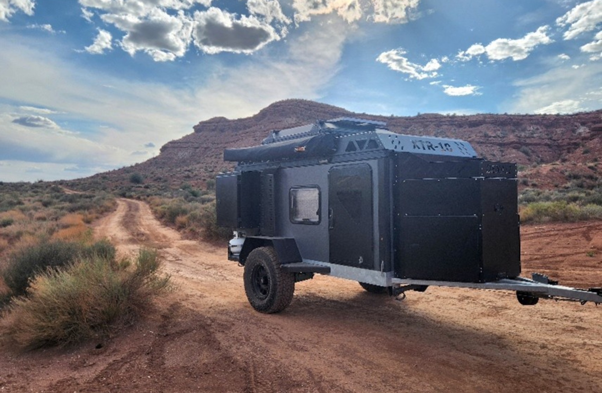 A rugged off-road camper trailer sits on a dirt path in a desert landscape with shrubs and rocky hills beneath a partly cloudy sky.