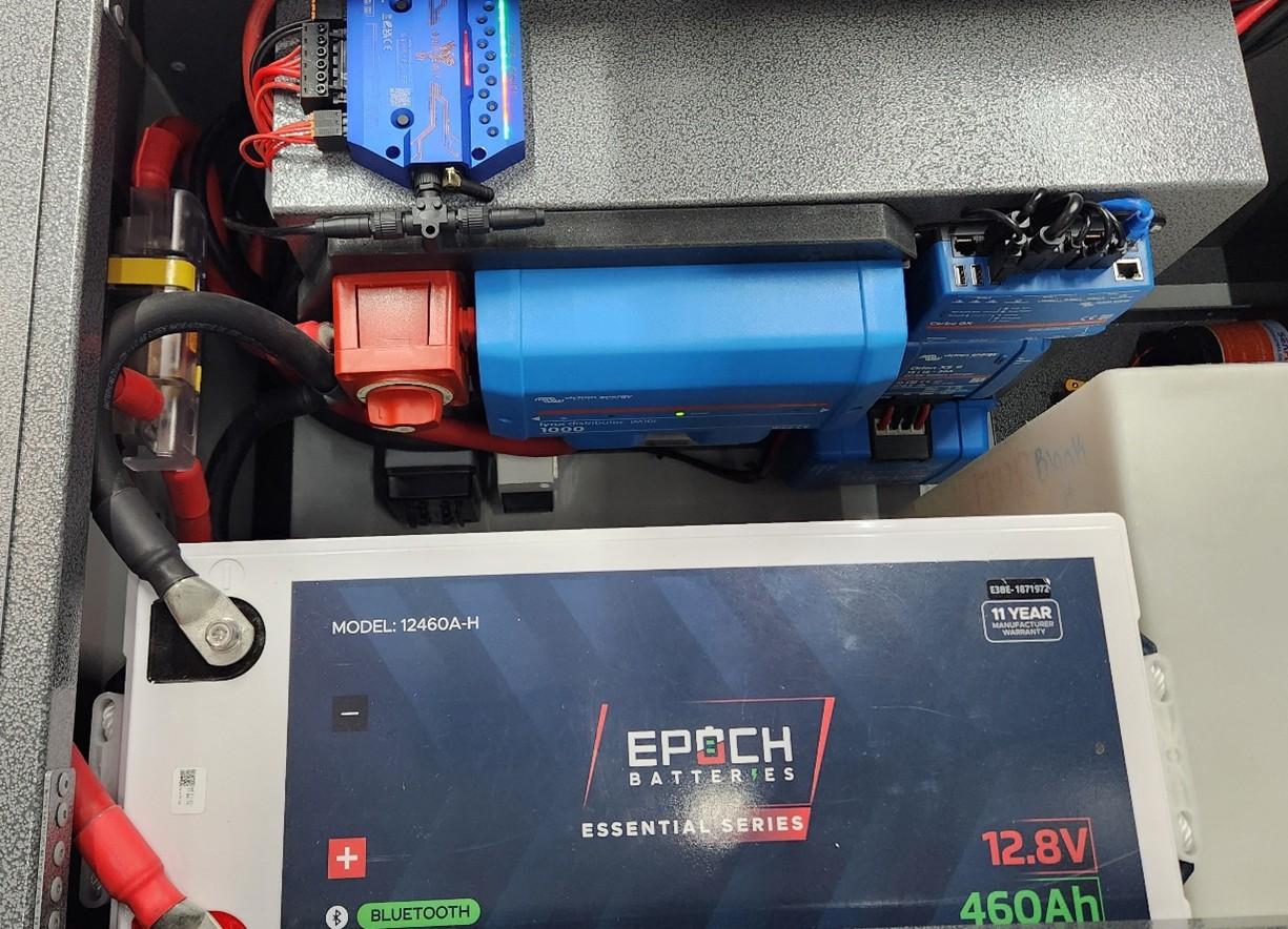 Close-up of an electrical setup featuring an EPOCH 12.8V 460Ah battery, a Victron inverter, switches, wiring, and various connected electronic components arranged in an organized manner.