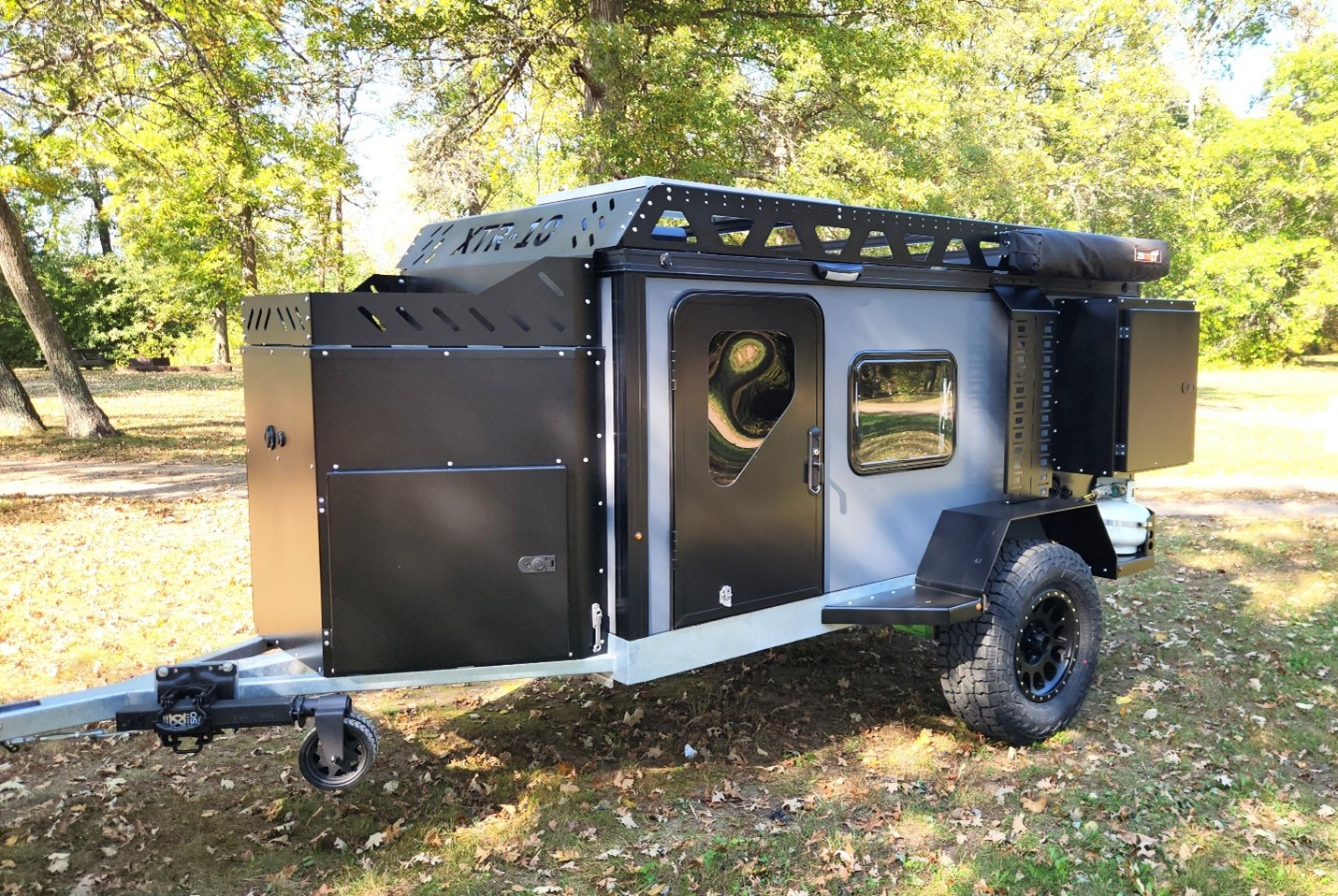 A rugged off-road camper trailer with large tires, black and gray panels, a side door with a round window, roof rack, and storage compartments—one of the sturdiest trailers parked on grass in a wooded area.