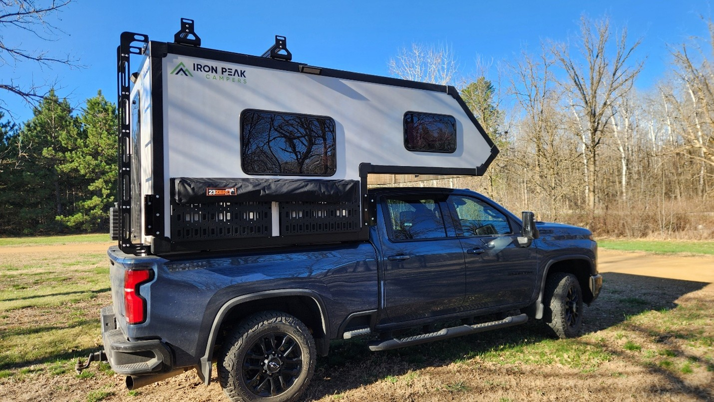 A dark gray pickup truck with an Iron Peak camper mounted on its bed is parked outdoors on a sunny day, surrounded by grass, trees, and a clear blue sky.