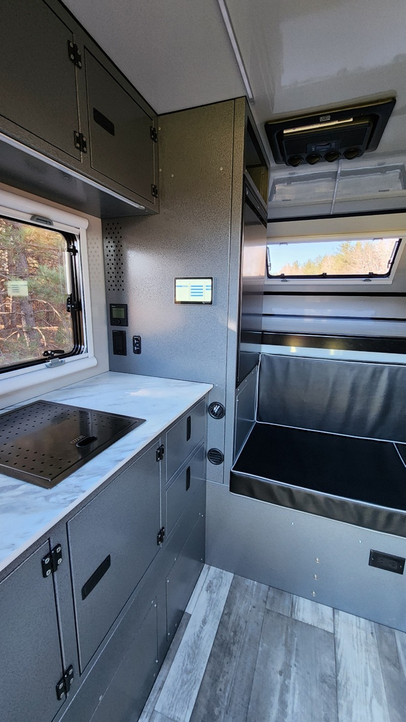 Interior of a modern camper van featuring a marble-look countertop with a built-in cooktop, gray cabinets, a window, control panel, and a seating or sleeping area by large windows. Wood-look flooring is visible.