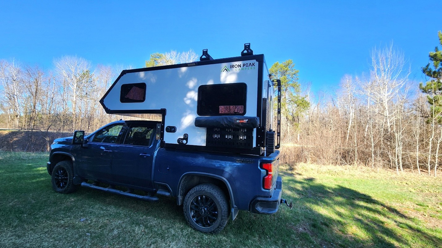 A dark blue pickup truck with a large white camper mounted on its bed is parked on grass near leafless trees and a clear blue sky—a perfect inspiration for building your camper adventure. The camper features black trim, windows, and visible equipment.