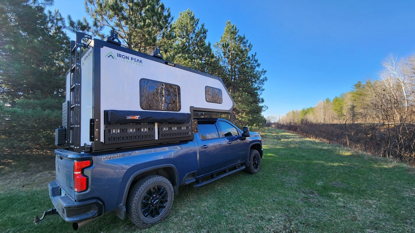 A blue pickup truck with an Iron Peak camper attached is parked on grass near a wooded area, under a clear blue sky—perfect inspiration if you want to build your camper for outdoor adventures.