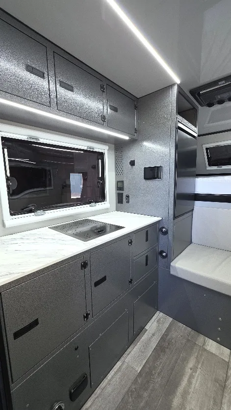 Modern camper van interior with gray cabinets, a marble-look countertop with a sink, large window, built-in appliances, overhead storage, a cushioned bench seat, and wood-look flooring. Bright, clean, and compact design.