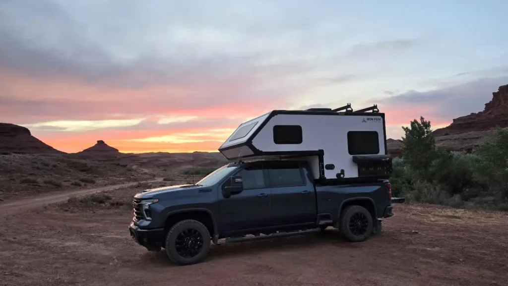 A dark pickup truck with a white camper attached is parked on a dirt road in a desert landscape at sunset, with colorful clouds and rocky formations in the background.