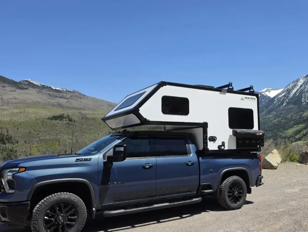 A dark blue pickup truck with an over-cab white camper is parked outdoors, surrounded by mountains and greenery under a clear blue sky.