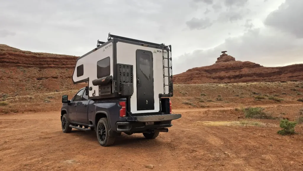 A black pickup truck with a white camper is parked on a dirt road in a desert landscape, with red rock formations and cloudy skies in the background.