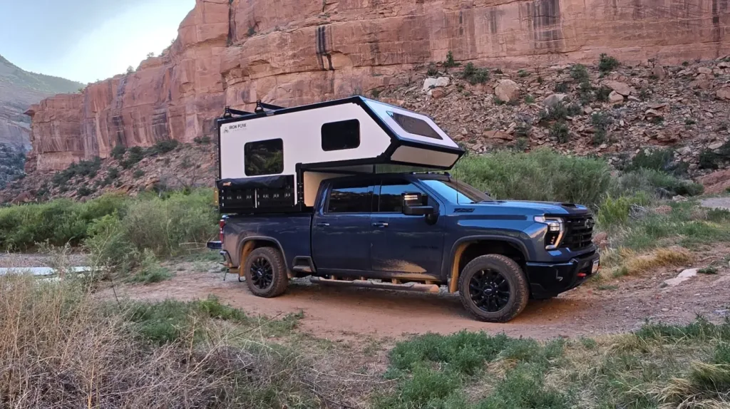 A blue pickup truck with a white and black camper attached is parked on a dirt road in a rugged, rocky canyon landscape with green bushes and tall red cliffs in the background.